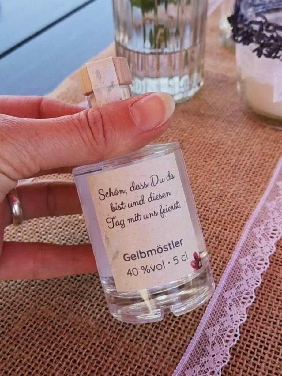Personalisiertes Gastgeschenk zur Hochzeit: 50ml Spirituosen-Mini mit verspieltem Etikett auf einer bunter Blumen-Tafel.